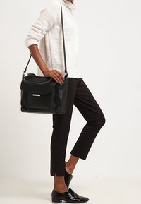 Sac bandoulière en cuir noir avec un rabat avant, une quincaillerie argentée et une finition texturée, associé à un pantalon noir et un pull blanc.