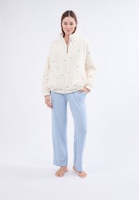 Futrzana kremowa kurtka z niebieskim kwiatowym haftem, dwoma kieszeniami i wysokim kołnierzem; w połączeniu z luźnymi, jasnoniebieskimi spodniami piżamowymi z mankietami.