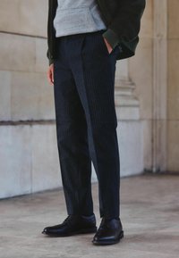 Jūras spēku režģa bikses, kas izgatavotas no teksturēta materiāla, apvienotas ar pulētu melnu ādas apavu pāri. Akcenti ietver sānu kabatas un piegriezumu, kas pielāgots.