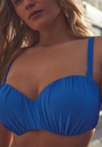 Gros plan d'une femme portant un haut de bikini bleu vif avec un tissu froncé et des bretelles fines, visible du torse au menton.
