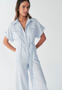 Mono de manga corta de color azul claro con botones en la parte delantera, dos bolsillos en el pecho, cintura con cinturón y un ajuste holgado. Hecho de tela suave y ligera.