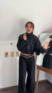 Giacca nera corta con motivo floreale, colletto alto e polsini elasticizzati abbinata a pantaloni neri a vita alta e una cintura marrone. Specchio sullo sfondo.