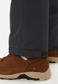 Pantaloni da esterno impermeabili grigio scuro con polsini in Velcro, abbinati a scarpe da trekking marroni con suole texturizzate e design supportivo.