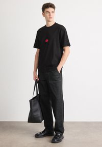 Black short-sleeve t-shirt with a red circle graphic, paired with dark denim pants and black loafers, holding a black tote bag.