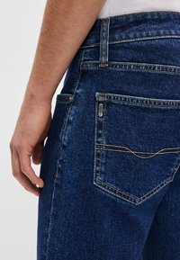 Tumesinised teksapüksid oranži õmblusega. Omadusteks on tagatasku ja sile tekstuur, näidatud on lähedalt vööpaneel.