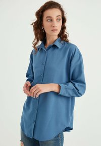 Camicia azzurra a maniche lunghe in morbido tessuto con bottoni frontali, colletto e orlo arrotondato. Caratterizzata da dettagli eleganti e vestibilità ampia.