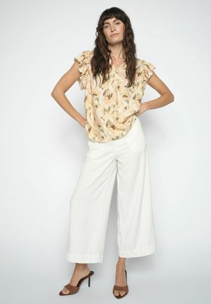 Žena s dlhými vlnitými hnedými vlasmi, oblečená v béžovej blúzke s volánikmi a krátkymi rukávmi s potlačou, bielych širokých nohaviciach a hnedých sandáloch na opätku.