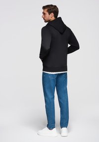 Felpa nera con cappuccio e vestibilità slim, abbinata a jeans azzurri chiari. Il modello si trova in piedi con le mani nelle tasche, mostrando la vista posteriore dell'outfit.