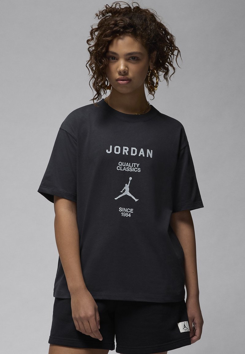 Black cotton t-shirt featuring white "JORDAN" text and a graphic logo of a jumping man. Short sleeves and relaxed fit. Paired with black shorts.