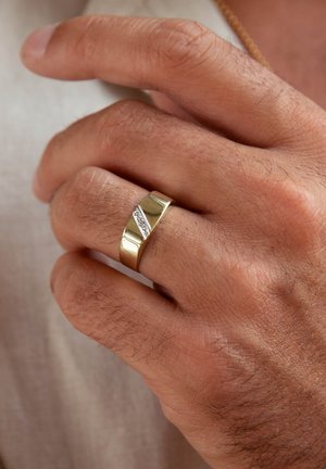 Gouden ring met een gepolijste afwerking, met een diagonale textuurband en een kleine accent van heldere stenen, liggend op een hand met licht haar.