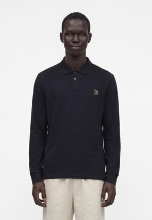 Uomo che indossa una polo nera a maniche lunghe con un piccolo logo ricamato con un elefante e pantaloni beige chiaro, in piedi davanti a uno sfondo bianco.