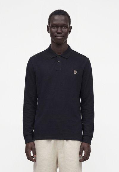Homme portant un polo noir à manches longues avec un petit logo éléphant brodé et un pantalon beige clair, debout devant un fond blanc.
