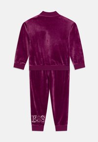 Tuta in velluto di un profondo viola, composto da una giacca con zip e pantaloni coordinati. I pantaloni presentano un dettaglio lucido "ESS" sull'anca.