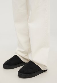 Pantofole in suede nero con suola in gomma piatta, abbinate a jeans color crema con cuciture visibili, che mostrano un fit rilassato.