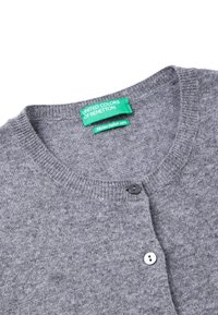 Šedý kardigan vyrobený z merino vlny. Má kulatý výstřih, vroubkované okraje a dva knoflíky – jeden černý a jeden bílý. Viditelné štítky.