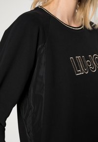 Musta huppari tyylikkäällä muotoilulla, jossa on kiiltävä musta sivupaneeli, kultareunus kauluksessa ja "LIU-JO" kultaisella brodeerattuna.