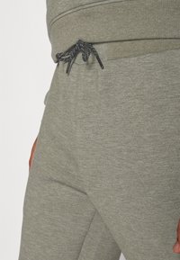 Sive trenerke napravljene od mekanog materijala s pojasom na vezanje, s crnim i srebrnim detaljima. Glatka tekstura i sužen kroj.