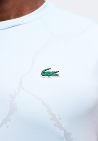 Logo crocodile vert brodé sur un tissu blanc texturé au niveau de la poitrine d'une chemise portée par une personne.