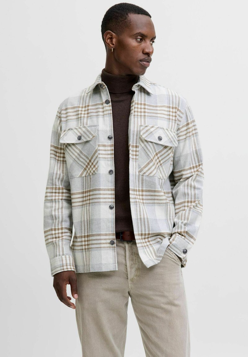 Camisa de cuadros en gris y beige, con botones en la parte delantera, dos bolsillos en el pecho y mangas largas. Lleva puesta una camiseta de cuello alto en marrón oscuro.