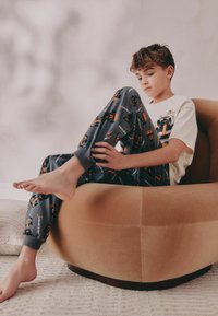 Grå pyjamasbukser med orange McLaren bilmønstre, parret med en beige t-shirt, båret mens jeg sidder på en rund brun stol.