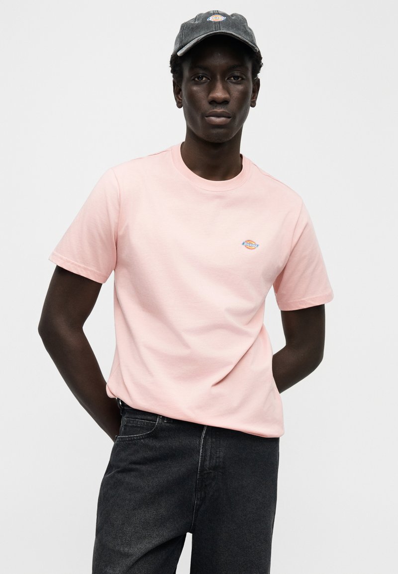 Lichtroze katoenen T-shirt met korte mouwen, ronde hals en een klein geborduurd logo op de borst. Gecombineerd met zwarte denim jeans en een pet.