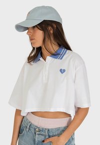 Polo shirt bianco a maniche corte con colletto blu e dettagli a righe, caratterizzato da un piccolo logo a cuore blu sul petto. Accompagnato da un berretto azzurro chiaro.