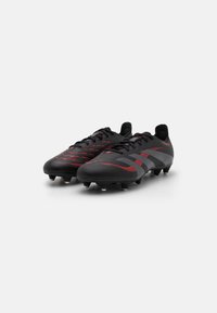 Botas de fútbol negras con acentos rojos, con una parte superior texturizada, un diseño elegante y múltiples tachuelas en la suela para tracción.