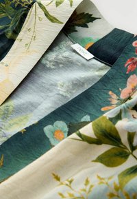 Floral patterned fabric showcasing vibrant flowers and leaves in shades of green, beige, and peach, with a visible brand label.
