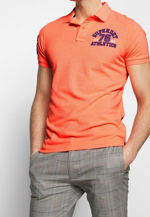 Homme portant un polo orange vif avec le logo bleu «Superdry 76 Athletics», associé à un pantalon à carreaux gris, mains dans les poches, sur fond blanc.