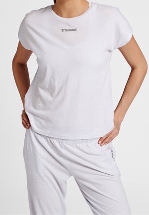 Kobieta ubrana w biały T-shirt z krótkim rękawem i dekoltem typu crew neck oraz pasujące białe spodnie z elastyczną talią, z rękami opartymi na biodrach.