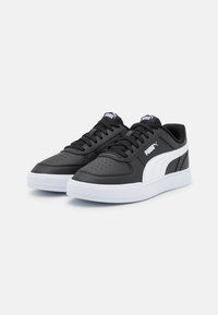 Mustat synteettisestä nahasta valmistetut tennarit valkoisilla yksityiskohdilla ja logolla. Varustettu rei'itetyllä kärjellä hengittävyyden lisäämiseksi ja litteällä kumipohjalla pidon parantamiseksi.