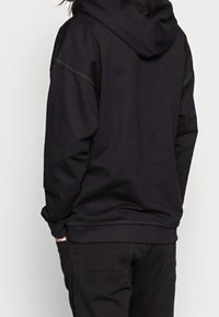 Felpa nera con cappuccio e cordino regolabile, texture morbida e dettagli di cucitura a contrasto. Indossata sopra pantaloni neri con una vestibilità comoda.