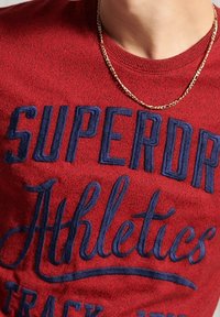 Camiseta roja de manga corta con cuello redondo que presenta el texto bordado en azul "SUPERDRY ATHLETICS" con un acabado texturizado. Se puede ver un collar de cadena dorada.