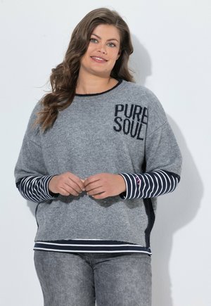 Vrouw met lang bruin haar die een grijze trui met de tekst "PURE SOUL" en een gestreept shirt met lange mouwen draagt, staand tegen een witte achtergrond.