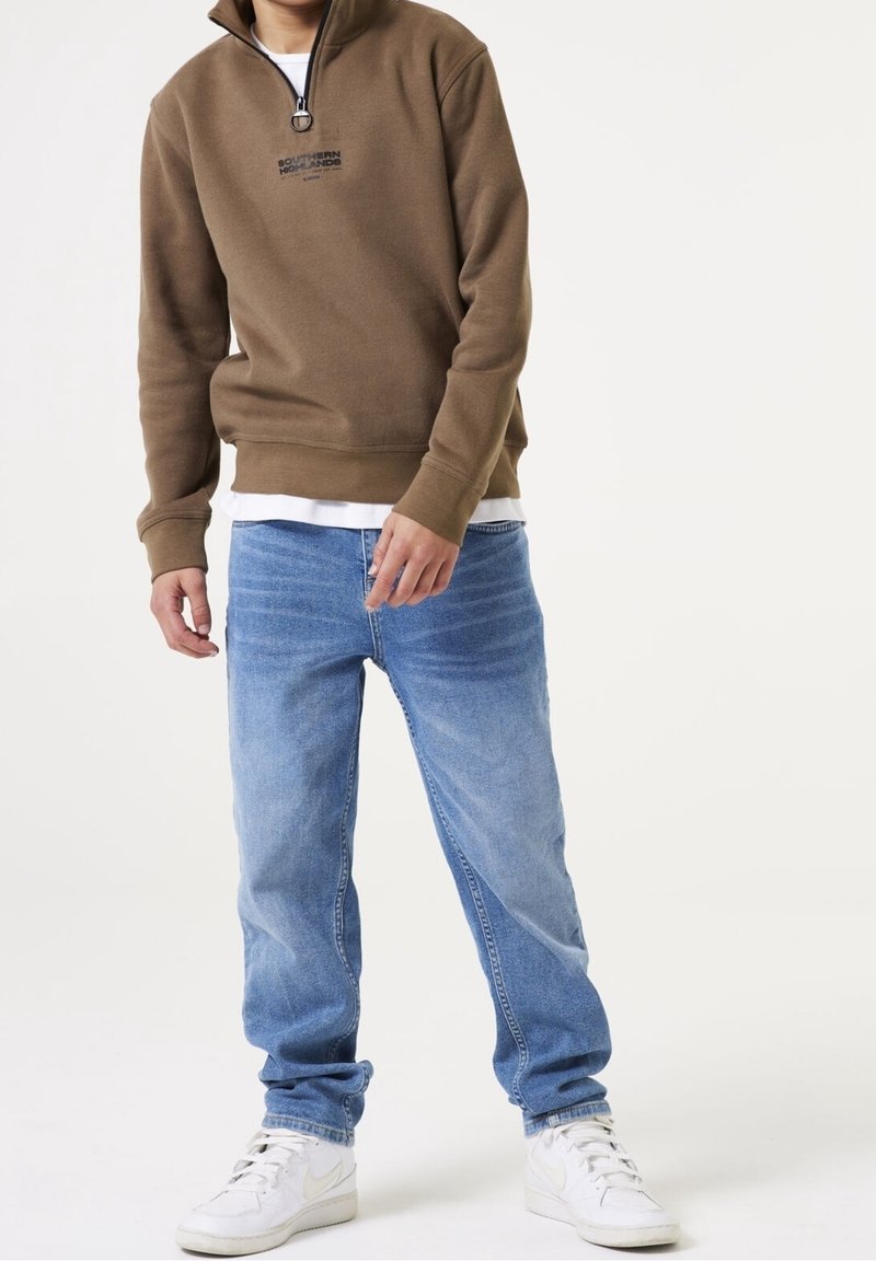 Sweat-shirt marron à fermeture éclair avec une doublure intérieure blanche, associé à un jean bleu clair et des baskets blanches. Coupe décontractée et silhouette relaxée.