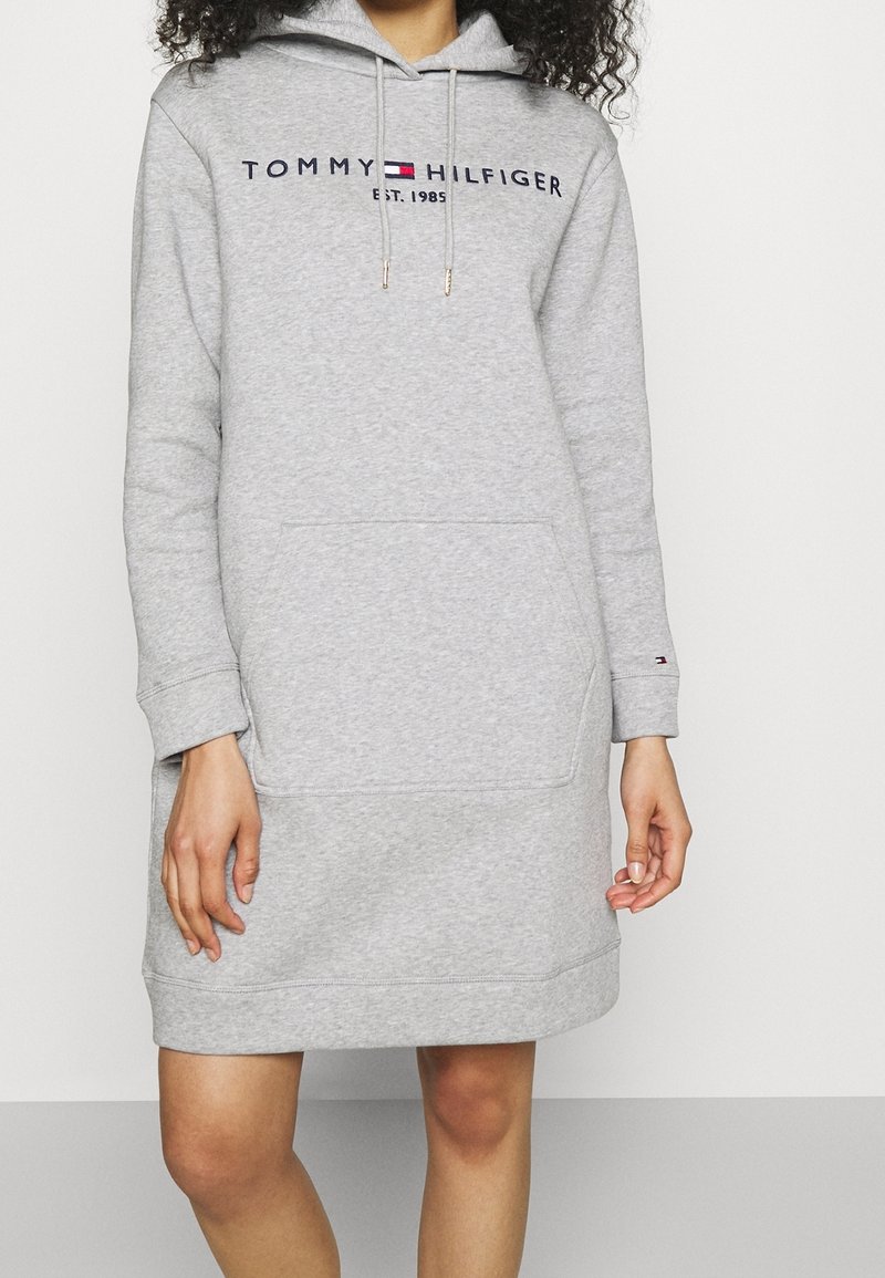 Femme portant une robe sweat à capuche Tommy Hilfiger gris clair avec une poche avant et un texte logo sur la poitrine.
