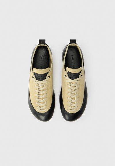 Parte superior de lienzo beige con punta y suela de goma negra, con cordones en contraste y un parche de cuero negro con marca en la lengua.