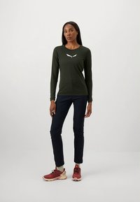 Pitkähihaisen tummanvihreän paidan v-muotoinen pääntie ja valkoinen logo, yhdistetty tummiin housuihin ja punaisiin lenkkareihin. Sileä kangas ja väljä istuvuus.