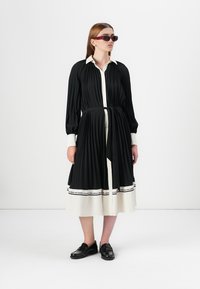 Black pleated dress with a white shirt collar and a belt, featuring a monochrome hem with text. Worn with black shoes and sunglasses.