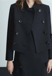 Veste noire à double boutonnage avec des boutons brillants, design structuré et col haut ; le matériau est lisse avec une silhouette ajustée et des coutures subtiles.