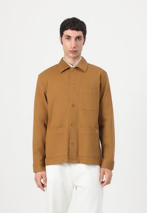Homme portant une veste marron à boutons avec trois poches avant par-dessus une chemise à carreaux et un pantalon blanc, debout devant un fond blanc.
