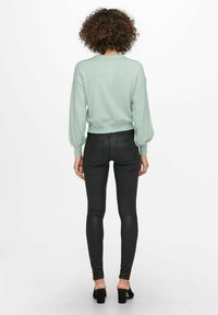 Mujer con pelo rizado de pie de espaldas, vistiendo un suéter verde claro, pantalones ajustados negros y zapatos de tacón negros, sobre un fondo blanco.