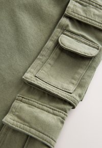 Gros plan sur un pantalon en tissu vert olive avec deux poches à rabat et des coutures détaillées sur la manche ou la jambe du pantalon.
