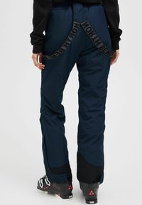 Navy blue ski pants with a textured surface, adjustable waistband featuring a black logo strap, and reinforced black cuffs at the ankles.