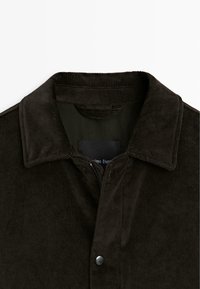 Giacca in corduroy marrone scuro con colletto a revore, chiusura a scatto e una fodera interna liscia. Tessuto testurizzato con leggere righe.