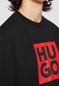 Czarny bawełniany t-shirt z dużą czerwoną prostokątną grafiką z wyraźnym czarnym napisem "HUGO". Okrągły dekolt i krótkie rękawy.