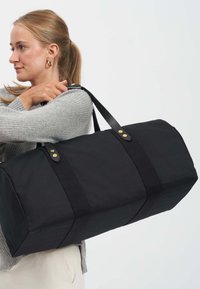 Černá plátěná taška přes rameno s dvojitými koženými uchy a zlatě tónovanými akcenty. Obdélníkový tvar s pevnou, strukturovanou vnější stranou.