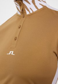 Brązowa koszulka polo z kołnierzykiem stojącym, zapięciem na guziki i białymi paskami. Na klatce piersiowej znajduje się logo w kolorze białym. Gładka tekstura.