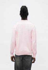 Ανοιχτό ροζ φούτερ με μακριά μανίκια, ribbed λαιμόκοψη και τελείωμα, λεία υφή, φορεμένο με σκούρο γκρι τζιν, απεικονίζεται από πίσω.