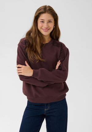 Une femme souriante aux longs cheveux bruns se tient les bras croisés. Elle porte un sweat-shirt couleur prune et un jean foncé, sur un fond clair et uni.